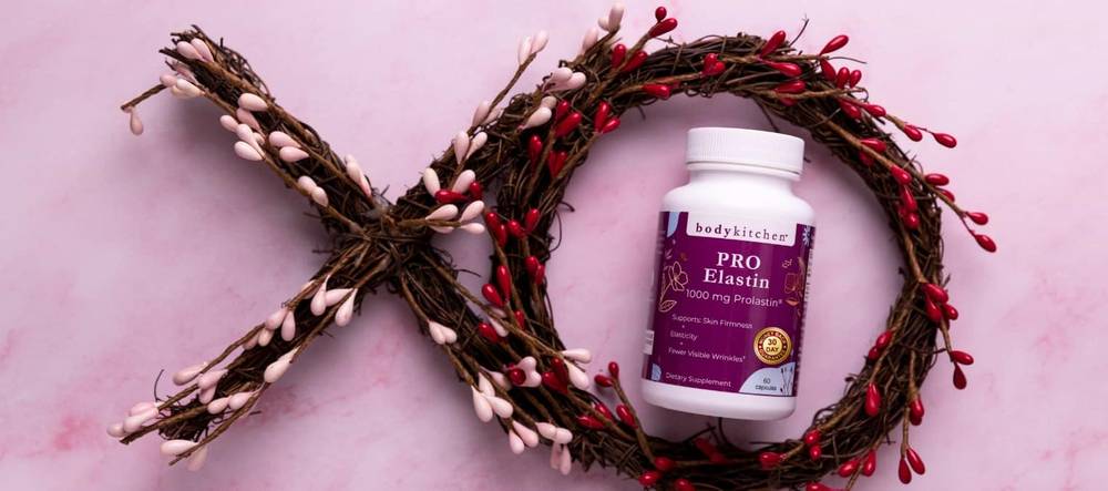 Bottle of Body Kitchen Pro-Elastin inside a twig wreath shaped like XO on a pink background
