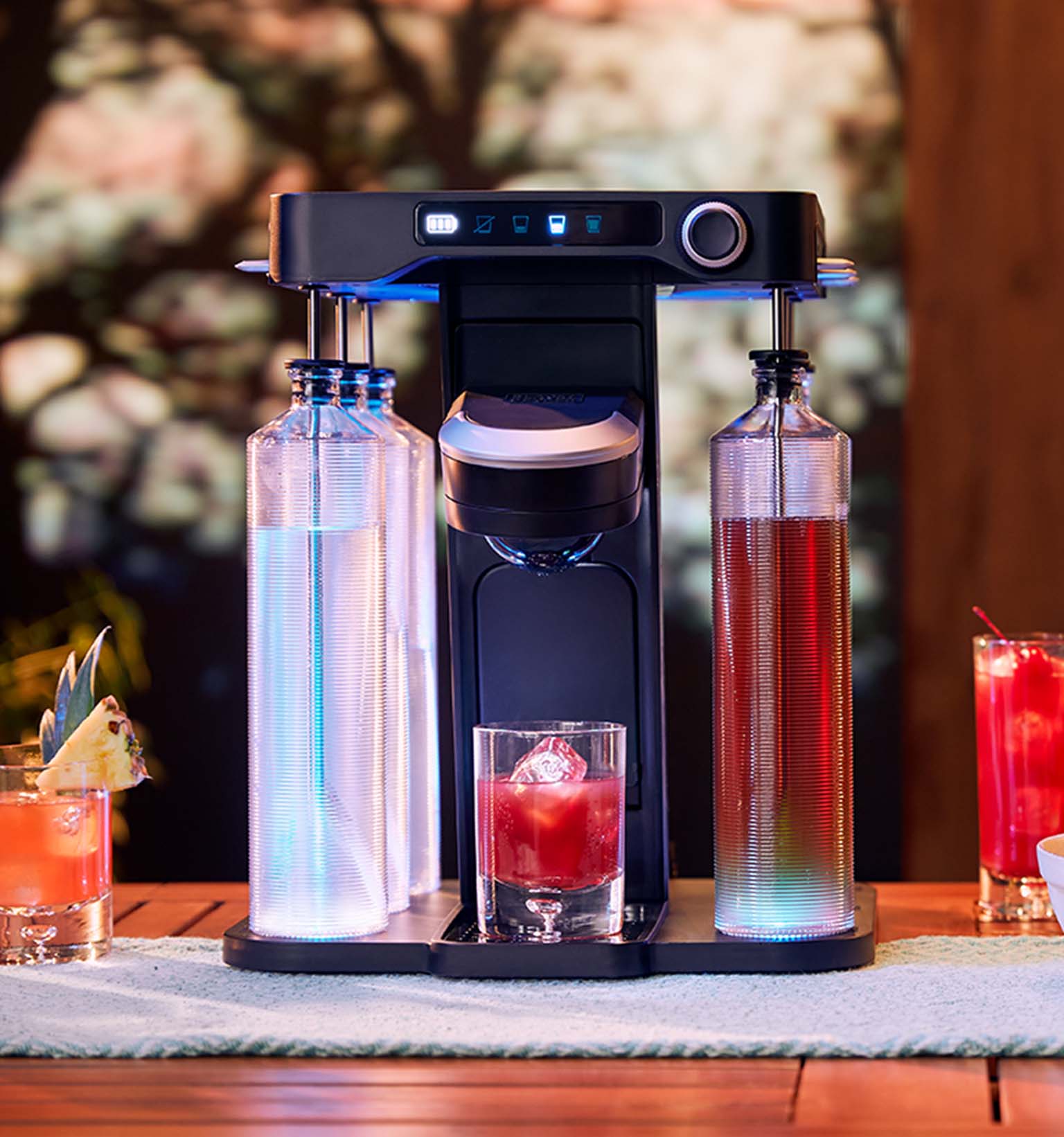 blue lights illuminated on the bev by BLACK+DECKER™ cordless cocktail maker sitting on a wooden patio table with drinks