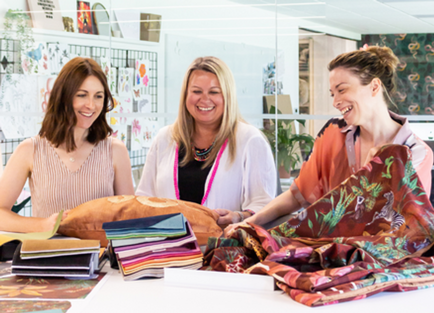 Three smiling people standing around a table of fabric samples and soft furnishings.
