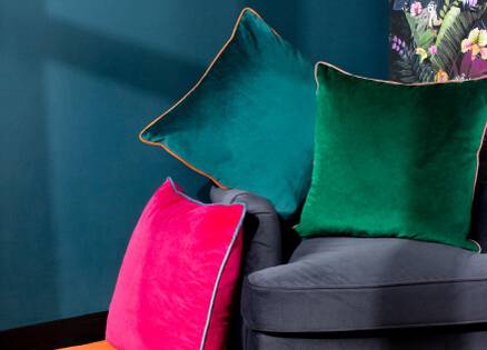 pink, teal and bottle green velvet cushion sitting on a grey chair with a blue painted wall