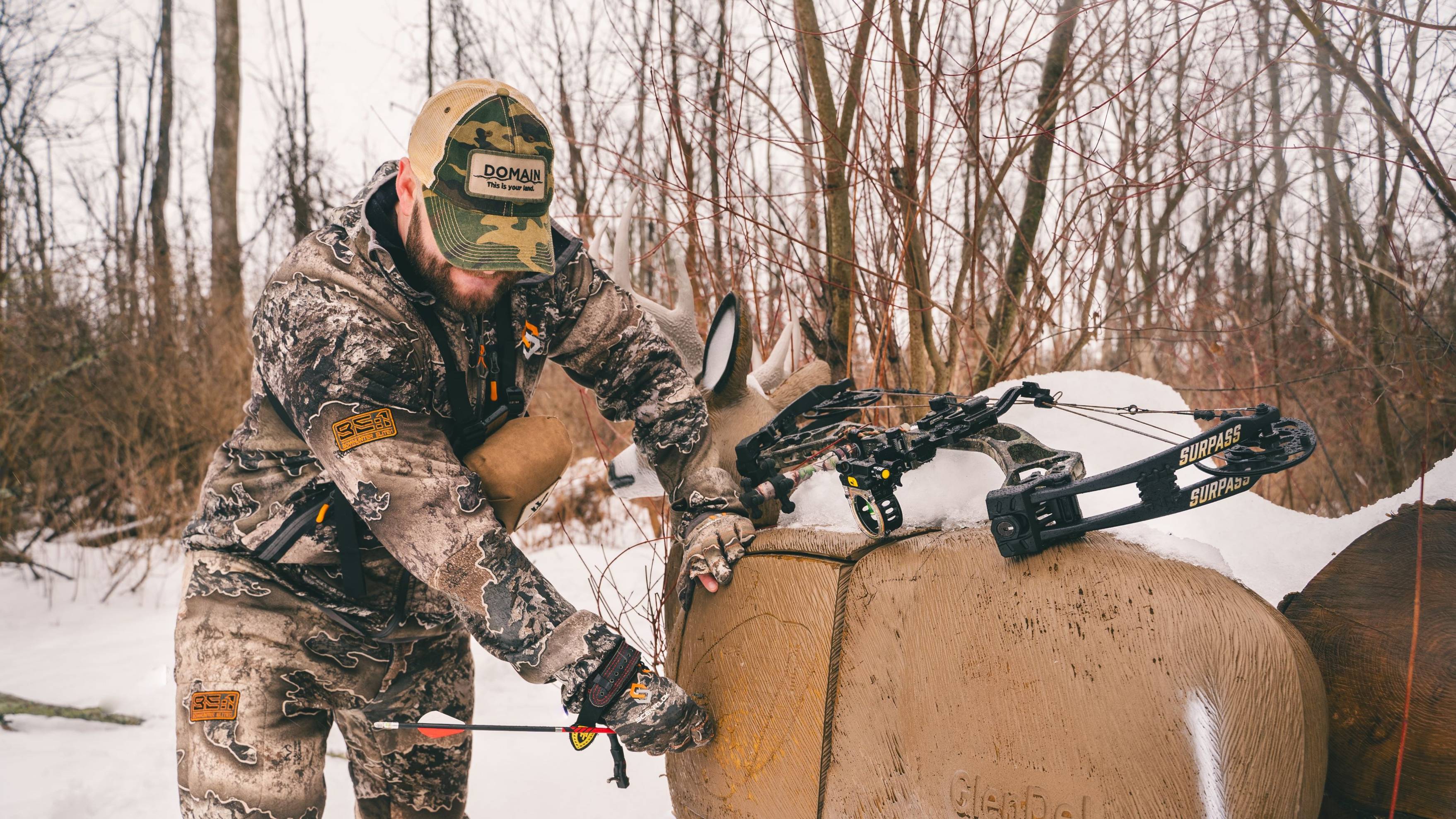 Man pulling arrows out of deer target with a Bear Archery Surpass Compound Bow laying on top in the snow
