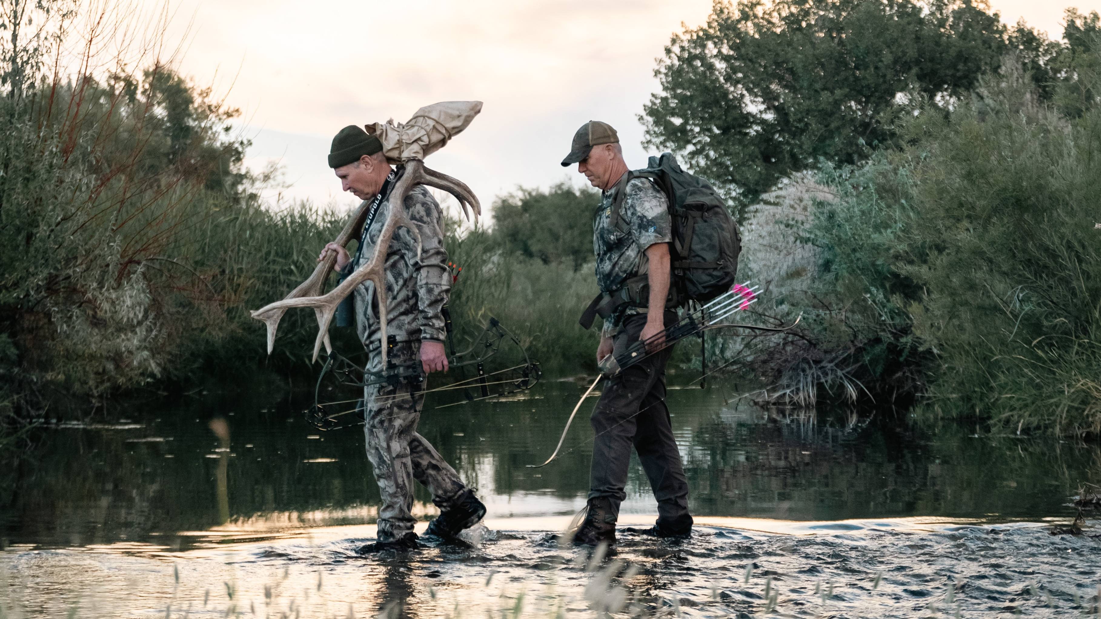 Chuck Adams and Fred Eichler carrying their Bear Archery bows across a creek. 