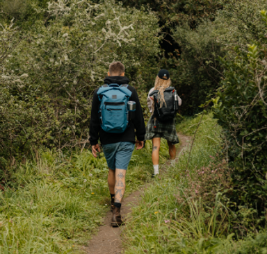 Woman carrying the Adventure Waterproof Backpack in Obsidian Black and Man carrying the Adventure Waterproof Backpack in Storm Blue along a footpath through a wood.