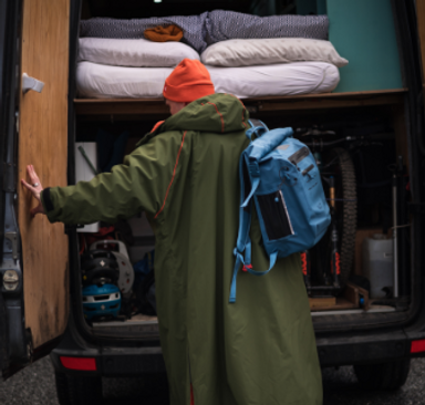 Woman wearing Red Equipment Voyager Beanie in Orange, Long Sleeve Pro Change Robe EVO in Parker Green and carrying the Adventure Waterproof Backpack in Storm Blue whilst closing the doors of a camper van.