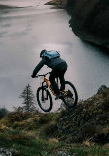 Man biking down mountain with a lake in the background. Carrying Red Equipment Adventure Waterproof Backpack in Storm Blue.