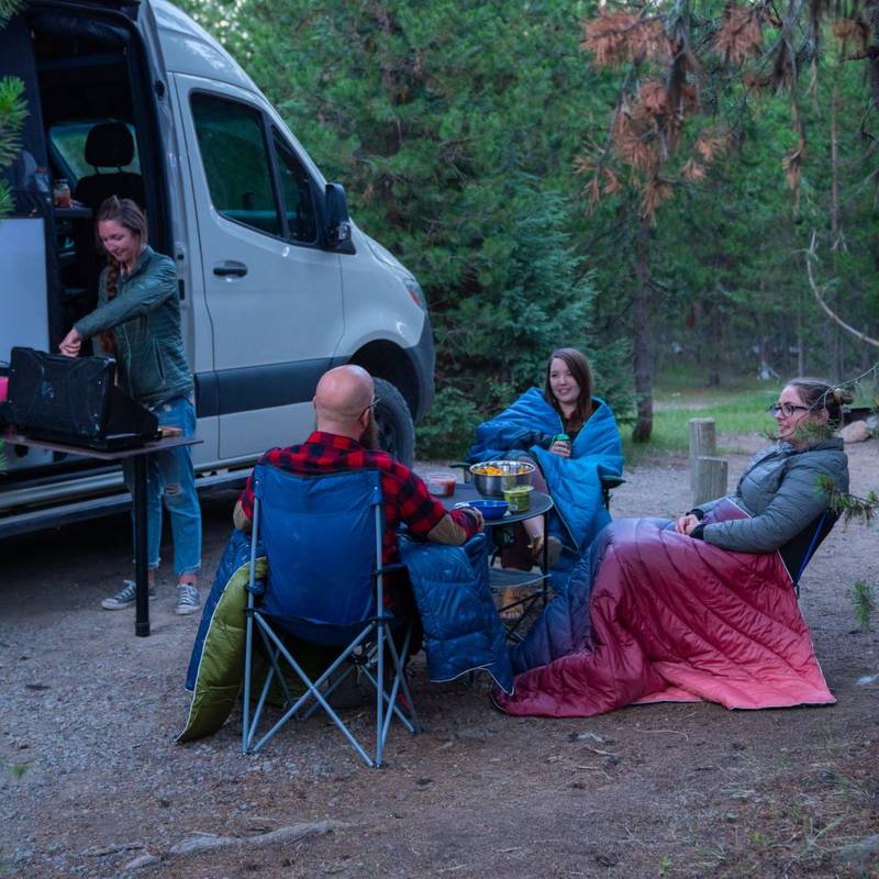 A group of friends relaxes outside their camper van, snuggled up in warm Rumpl camper van blankets while enjoying each other's company and the beautiful scenery.