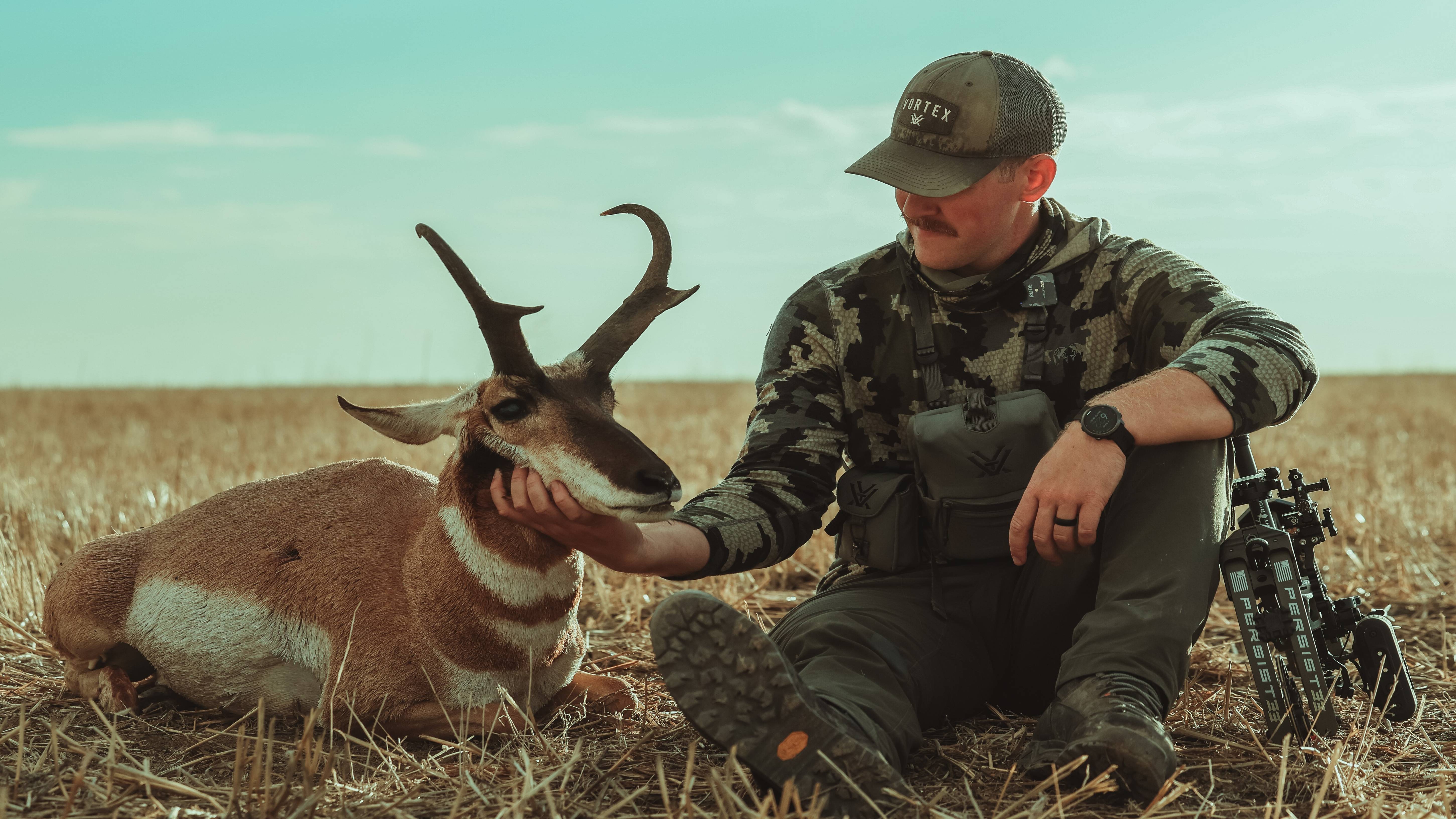 Man resting against Bear Archery Persist 33 compound bow in a field with an antelope.