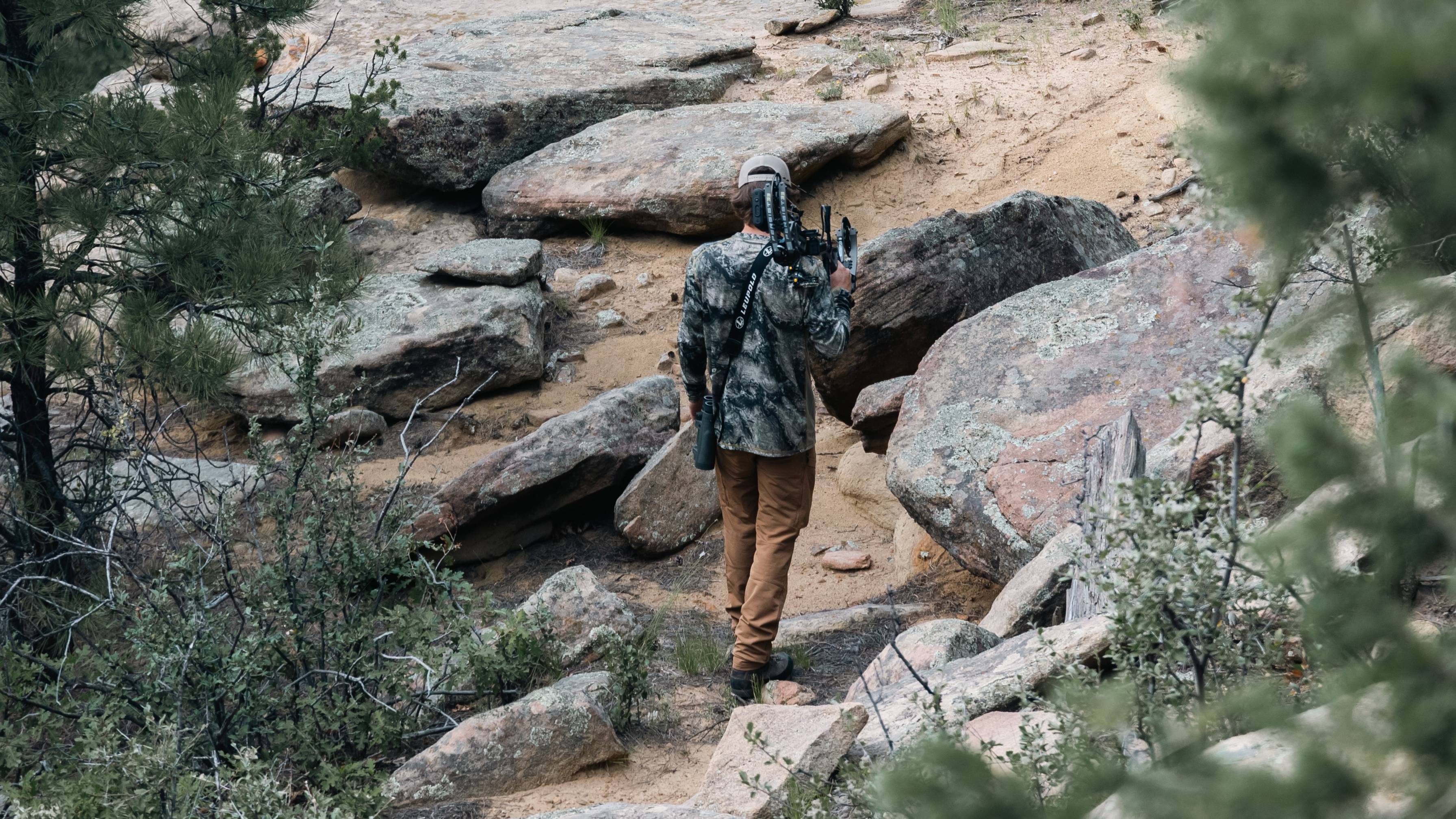 Man holding a Bear Archery Persist 33 compound bow over his shoulder as he walks up a mountain.