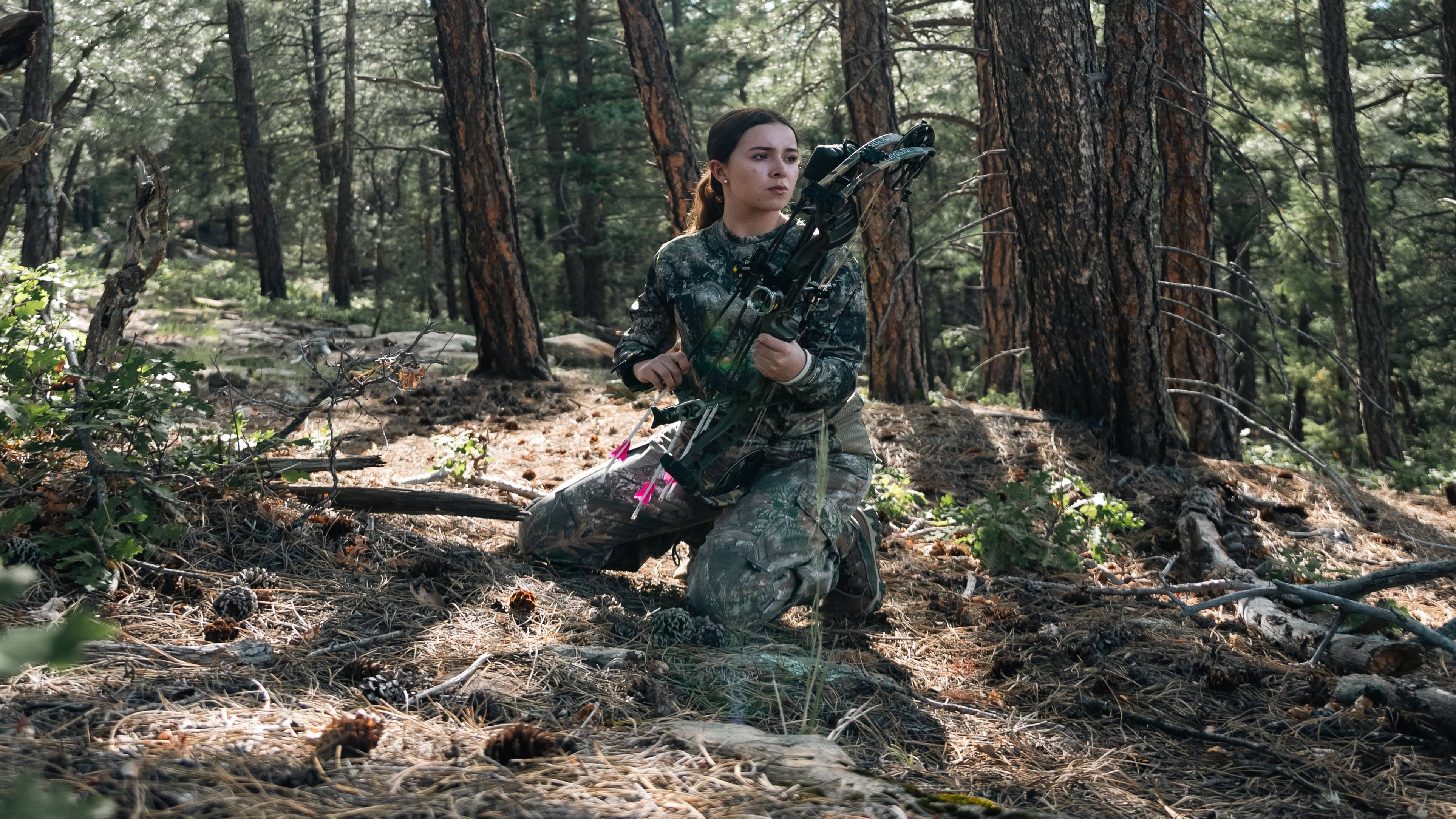 Girl surrounded by trees, focused on the distance while holding a Bear Archery Legend 30 RTH Compound Bow