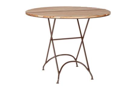 The nkuku Vivika Outdoor Dining Table - Natural sits on a stone patio, paired with two matching metal chairs with wooden seats. Stacked plates and napkins rest on the table, while lush greenery and white flowers fill the garden backdrop.