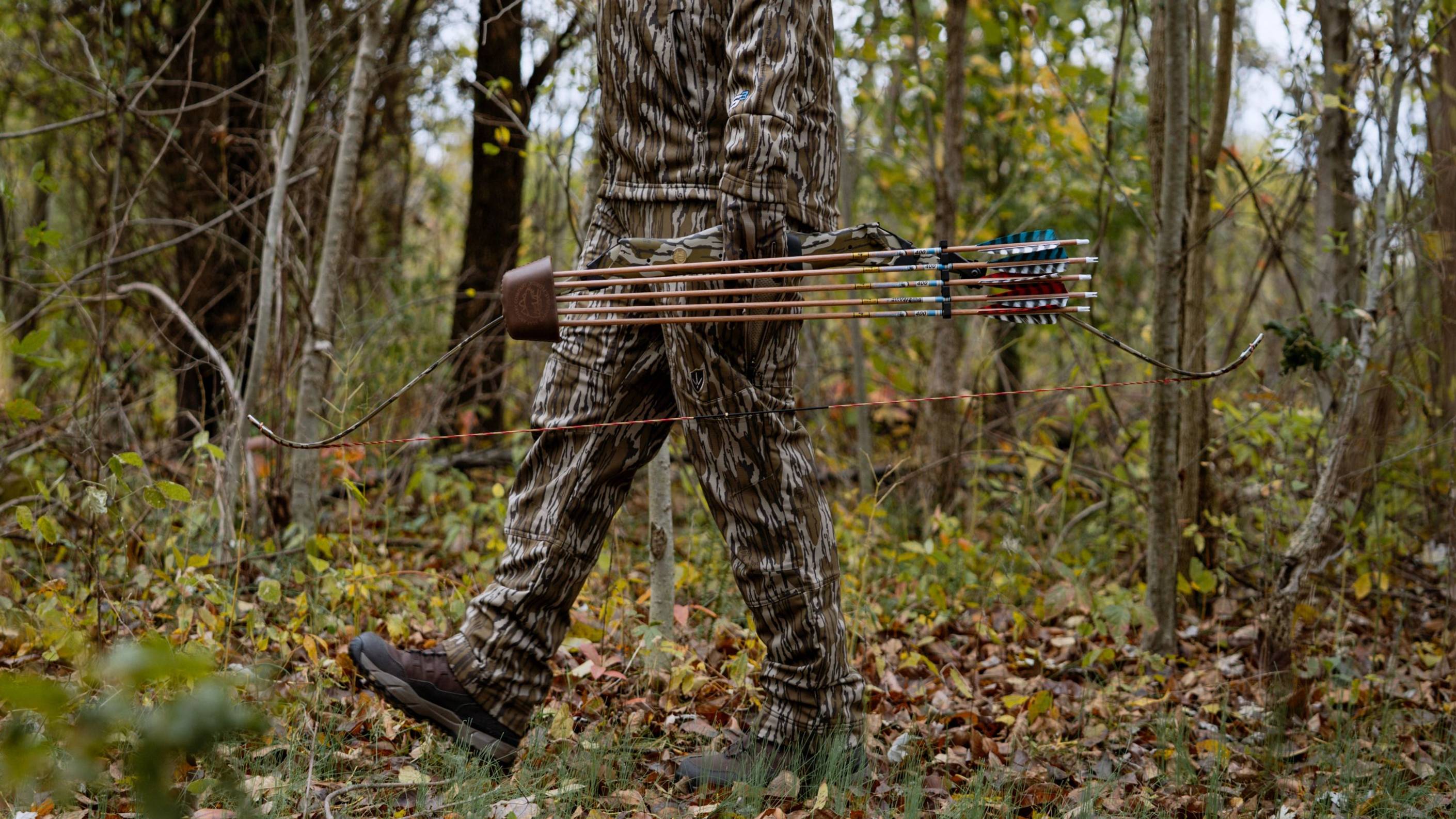 Person walking through woods with takedown recurve bow with Mossy Oak Bottomland Camo Take Down Bow Limbs from Bear Archery.