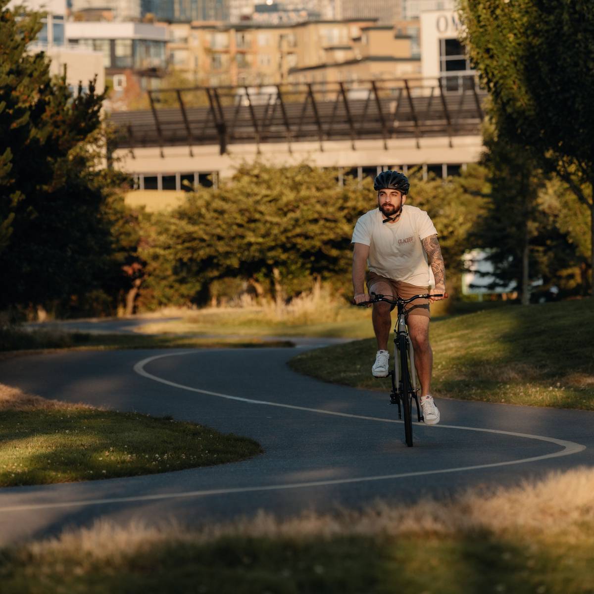 A cyclist wearing a Silas Bike Helmet and white shirt riding on a bike trail with a city landscape in the background.