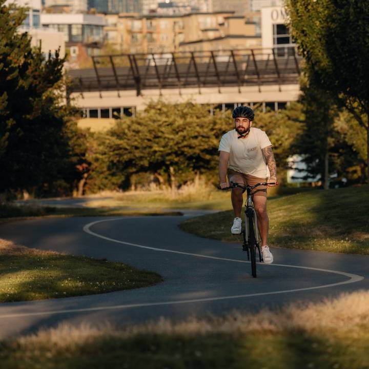 A cyclist wearing a Silas Bike Helmet and white shirt riding on a bike trail with a city landscape in the background.