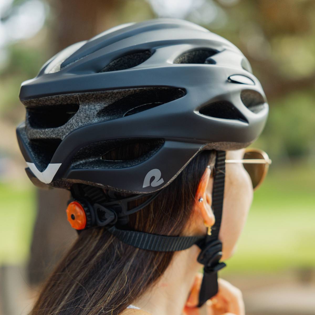 A person wearing a black Silas Road Bike Helmet with LED light at the rear shown with long straight hair peeking out and a green blurred background.