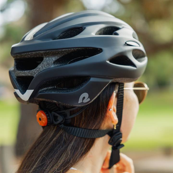 A person wearing a black Silas Road Bike Helmet with LED light at the rear shown with long straight hair peeking out and a green blurred background.