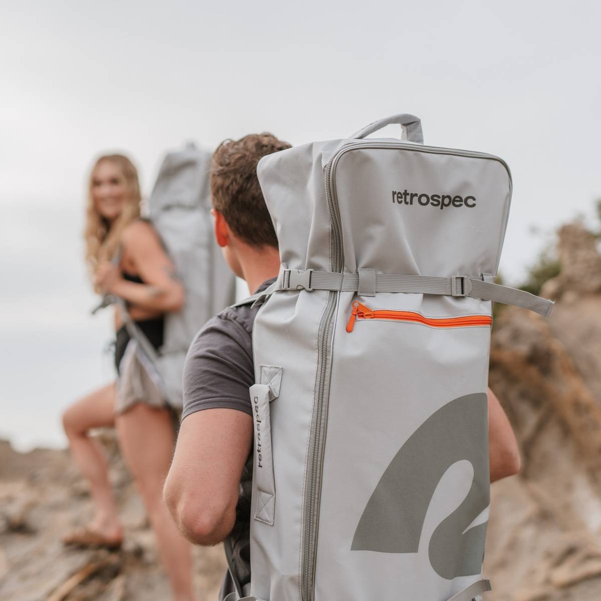 Two hikers ascend a rocky terrain, each carrying a large, gray retrospec backpack with an orange zipper detail.