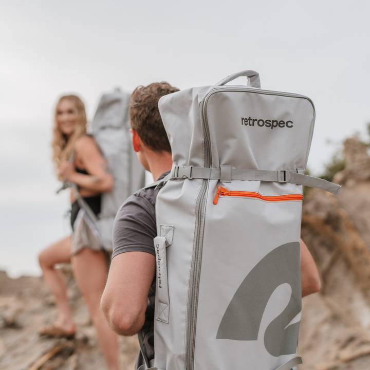 Two hikers ascend a rocky terrain, each carrying a large, gray retrospec backpack with an orange zipper detail.