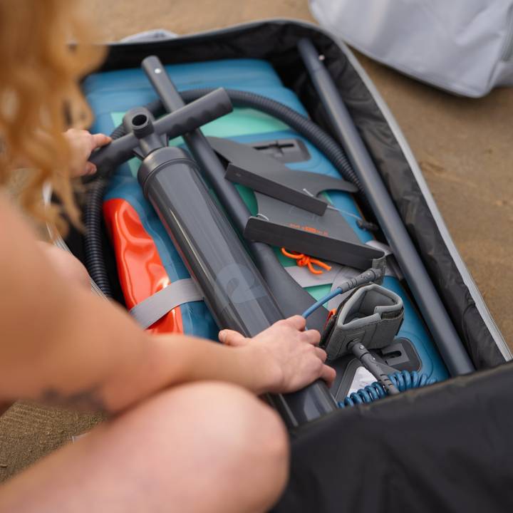 A person organizes a compact backpack filled with a pump, inflatable gear, and various accessories for a water activity.