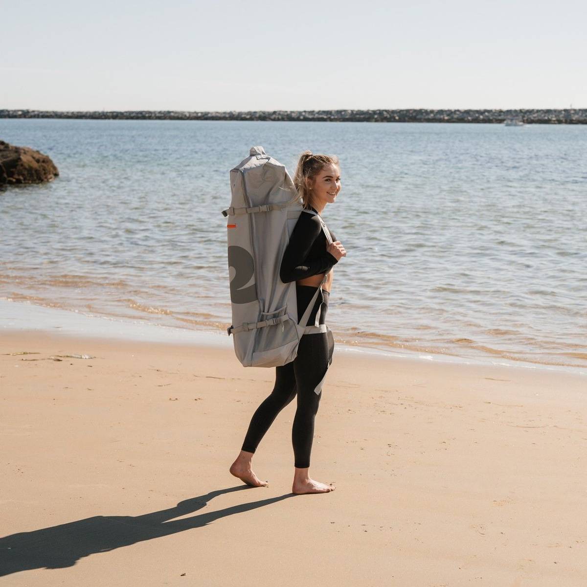 A person in a black wetsuit walks along a sandy beach carrying a large grey backpack, with calm water and a clear sky in the background.