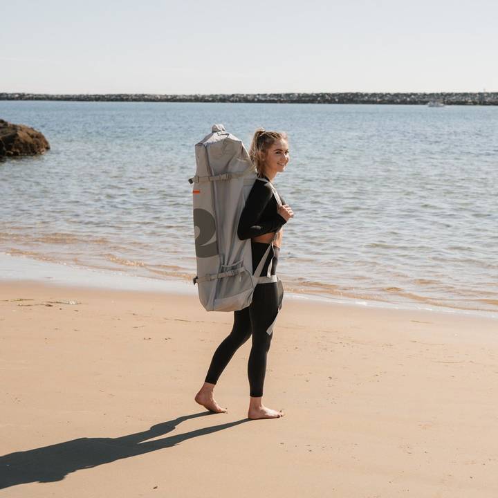 A person in a black wetsuit walks along a sandy beach carrying a large grey backpack, with calm water and a clear sky in the background.