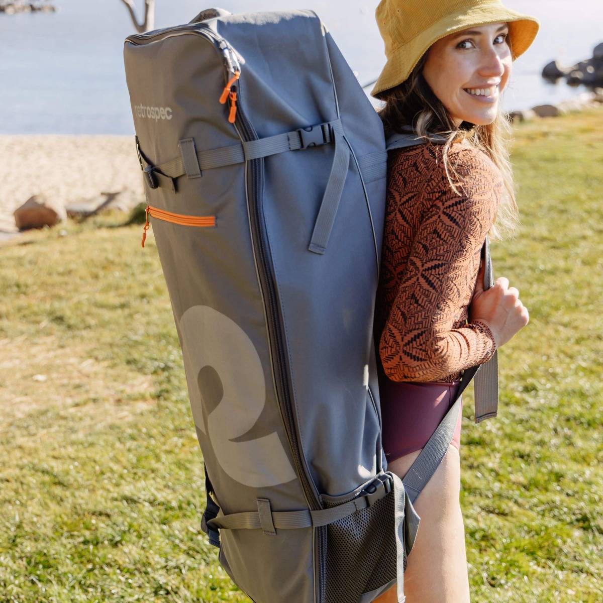 A person stands on the beach, wearing a patterned top and hat, carrying a large gray backpack with an orange zipper and logo.