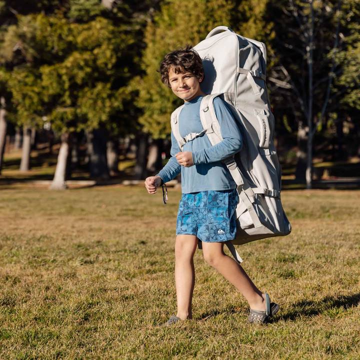 A child walks across a grassy area, wearing a blue long-sleeve shirt and patterned shorts, carrying a large gray backpack.