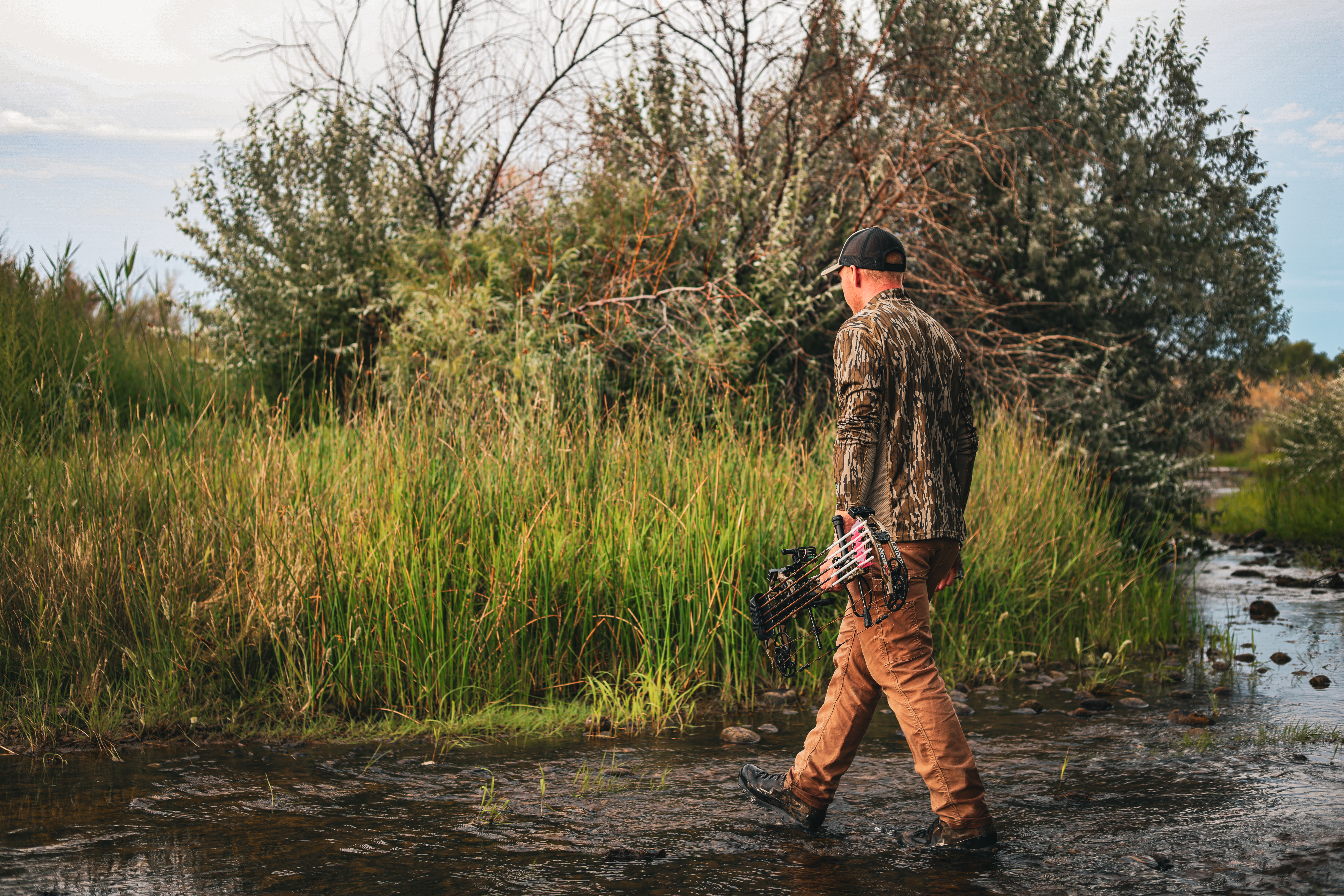 Man walking across a stream holding the Bear Archery Legit Pro compound bow.