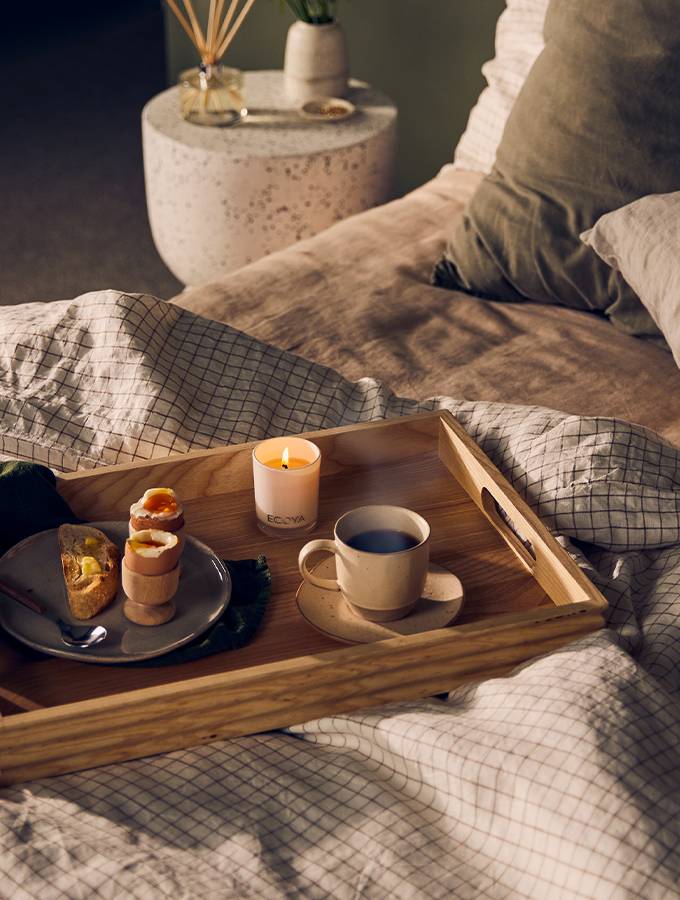Cozy breakfast-in-bed scene with a wooden tray holding a lit ECOYA candle, soft-boiled eggs, toast, and a ceramic cup of coffee, styled on a neutral grid-patterned duvet with warm-toned bedding and a terrazzo bedside table in the background