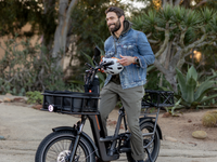 A man smiling while standing over the RadRunner 3 Plus. He is holding a helmet & his bike is outfitted with a large front & back basket. 