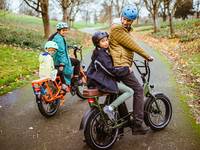 A woman of color rides an orange RadWagon with her daughter on the back. A man rides a green RadRunner with a child on the back seat.