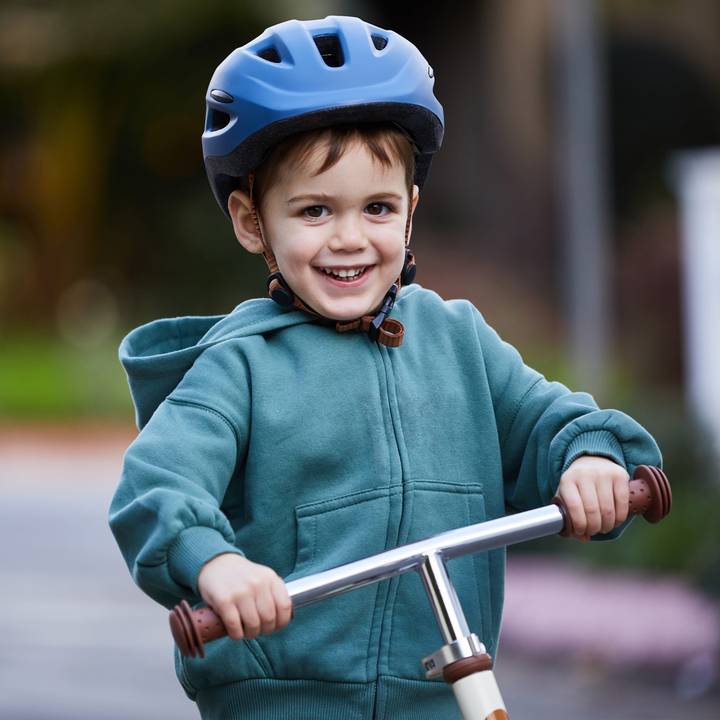 A young child wearing a blue bicycle helmet and a green hoodie enjoys riding a scooter outdoors, gripping the handlebars tightly.