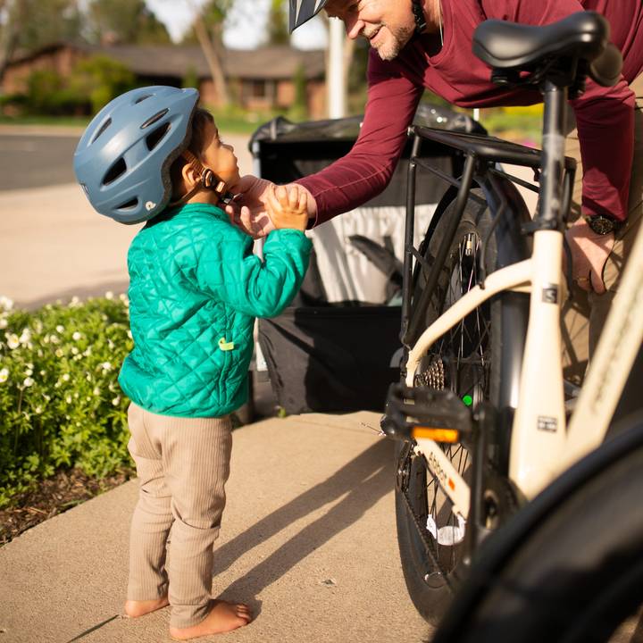 A young child in a green quilted jacket interacts with a person near a bicycle, showcasing a warm and playful moment outdoors.