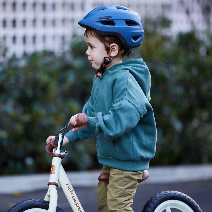A young child wearing a blue bicycle helmet and a green hoodie enjoys riding a bike outdoors, gripping the handlebars tightly.