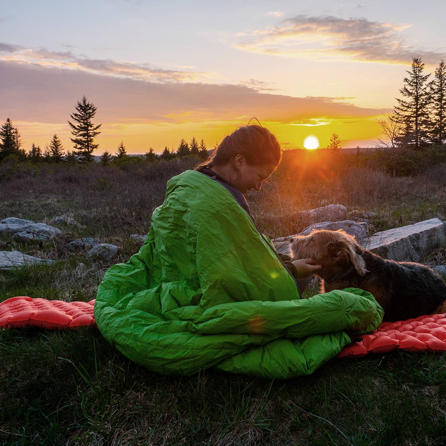 A person wrapped in a green sleeping bag sits on a red sleeping pad, enjoying a sunset in a natural setting with trees in the background. A dog is beside them.