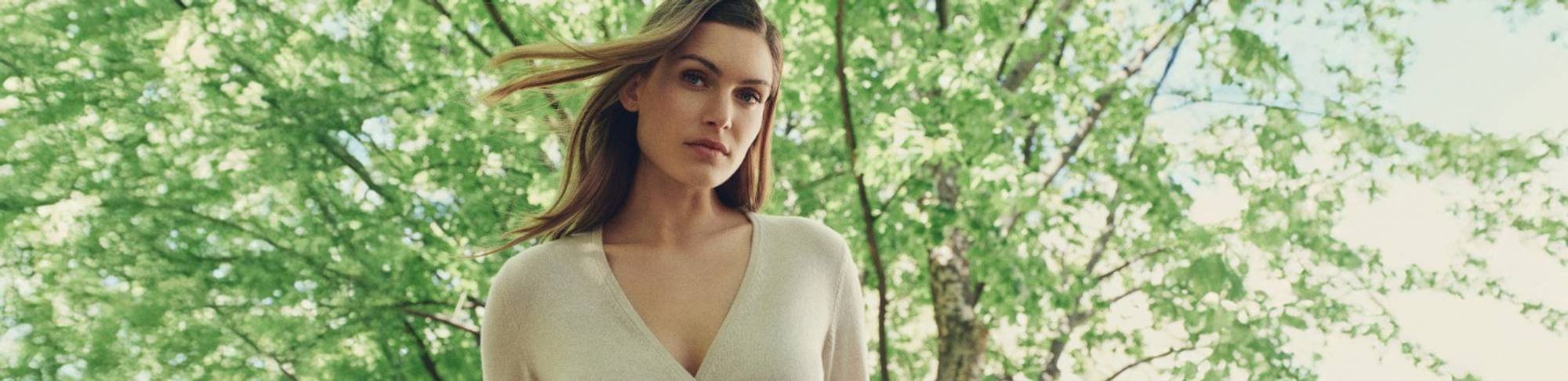 Woman wearing a light wrap-front top, standing outdoors with green leafy trees in the background and sunlight filtering through