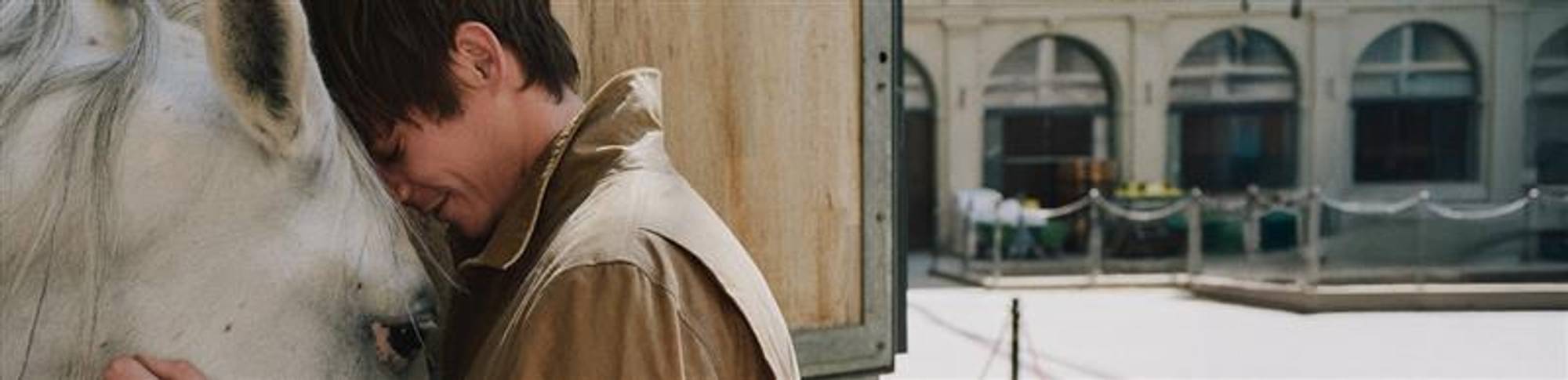 A man wearing a brown jacket gently leans his forehead against a white horse's forehead, sharing a quiet moment of connection. They are standing outdoors near a wooden structure, with a building and paved courtyard visible in the background