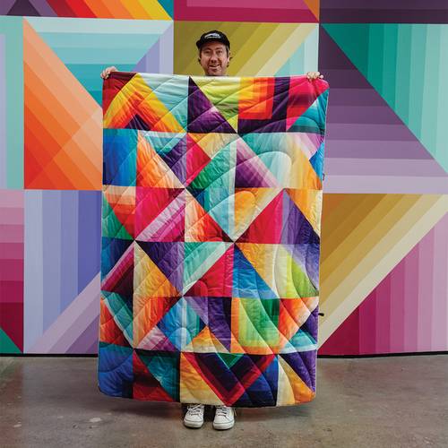 Nathan Brown holding up the Original Puffy Blanket - Cozy Dimensions in front of the Cozy Dimension mural at the Rumpl HQ in Oregon.