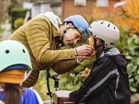 A light-skinned man wearing a helmet helps to clips his daughter's helmet before they go for a Rad ride.
