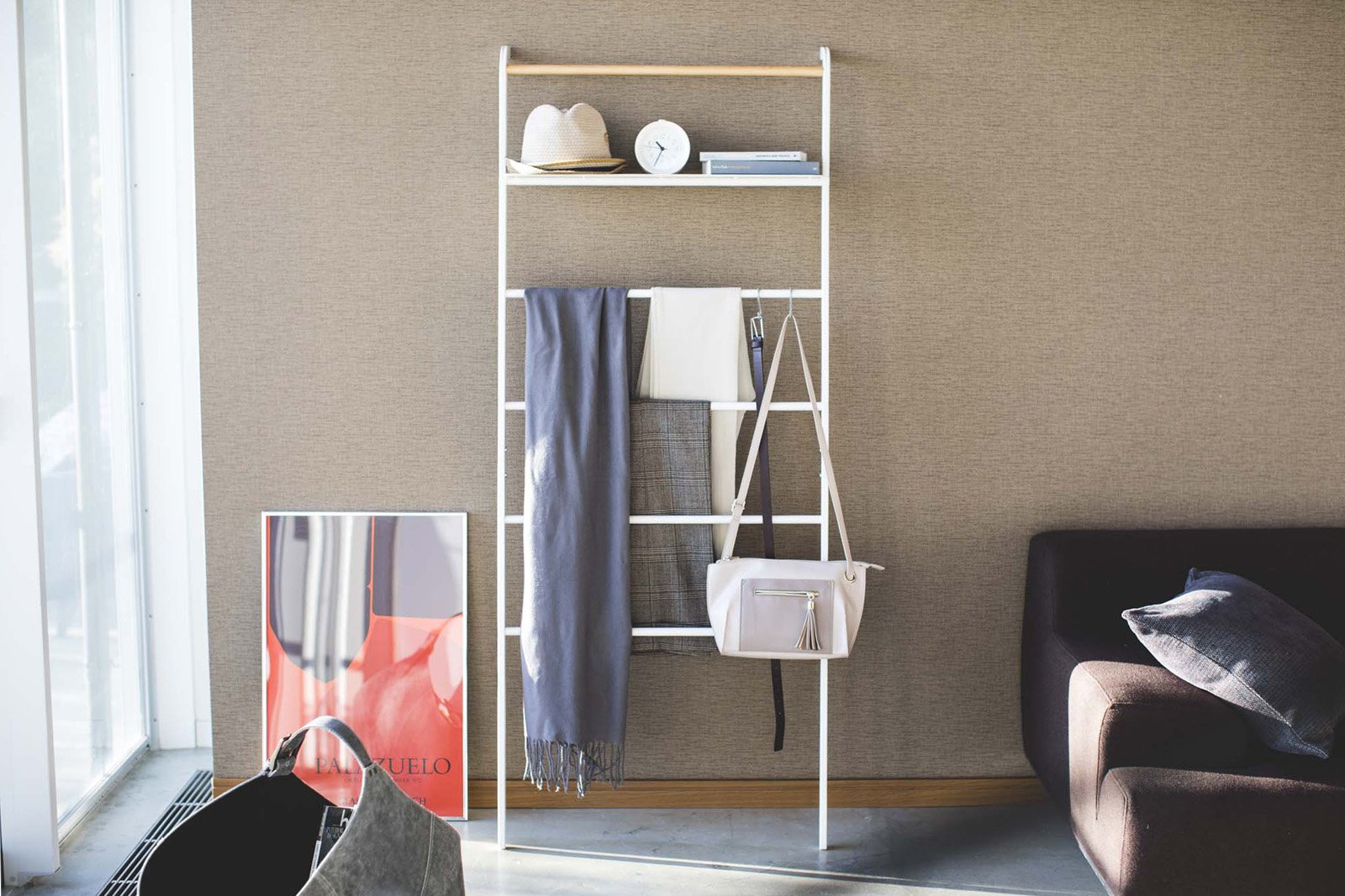 Yamazaki Home white Leaning Ladder Rack with Shelf holding a scarf, a hat next to a sofa. 