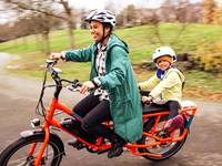 A woman of color smiles as she rides her orange RadWagon, with her daughter on the back of the electric bike, holding the Rad Power Bikes Deck Hand.