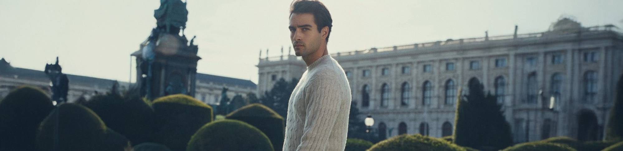 Man wearing a light cable-knit sweater, standing in a manicured garden with a grand historic building and statue in the background
