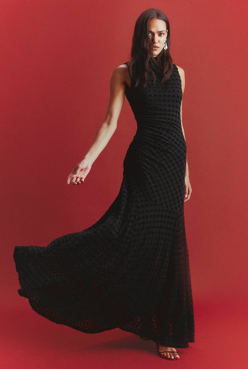 A woman in a sleeveless black gown with a textured gingham pattern and a flowing skirt poses against a red backdrop, wearing drop earrings and strappy black heels.
