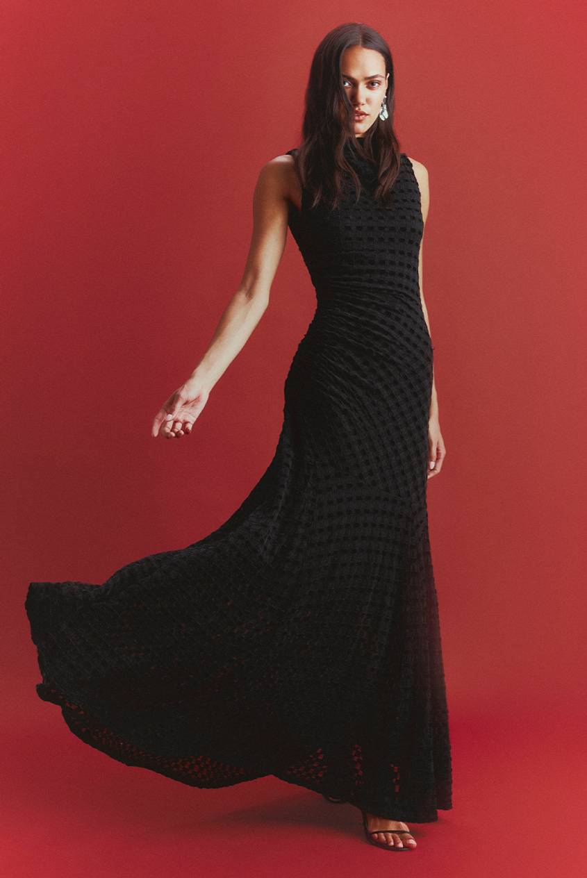 A woman in a sleeveless black gown with a textured gingham pattern and a flowing skirt poses against a red backdrop, wearing drop earrings and strappy black heels.