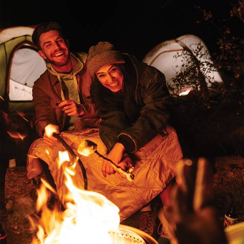a man and woman laughing while roasting marshmallows over a campfire, a NanoLoft® Flame Blanket is spread out over both their legs