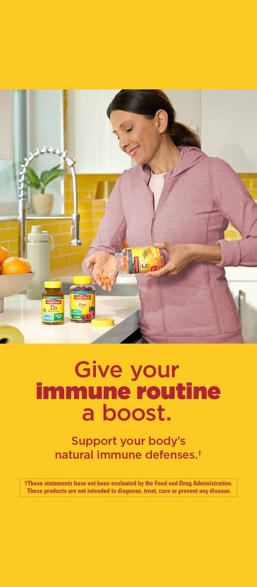 Person holding a bottle of Nature Made immune support supplements, with other vitamin bottles and fresh oranges on the counter. Text: ‘Give your immune routine a boost. Support your body’s natural immune defenses.’