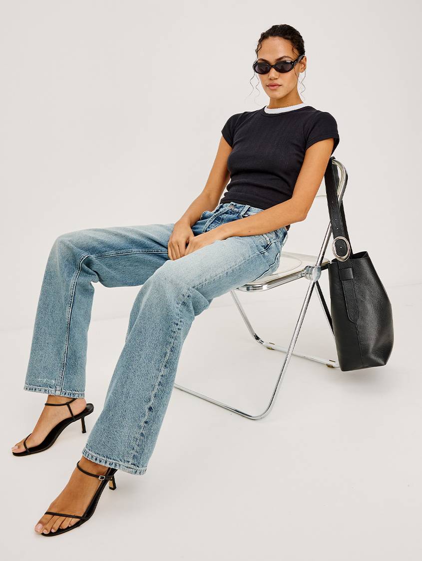 Media Card of the Del Mar Boyfriend Jean in Vintage Azure, Black Short Sleeve Tee with a White Collar, and Torino Bucket Bag in Black Pebble Leather. Full Body Front Angle View Featuring Model Sitting in a Chair.
