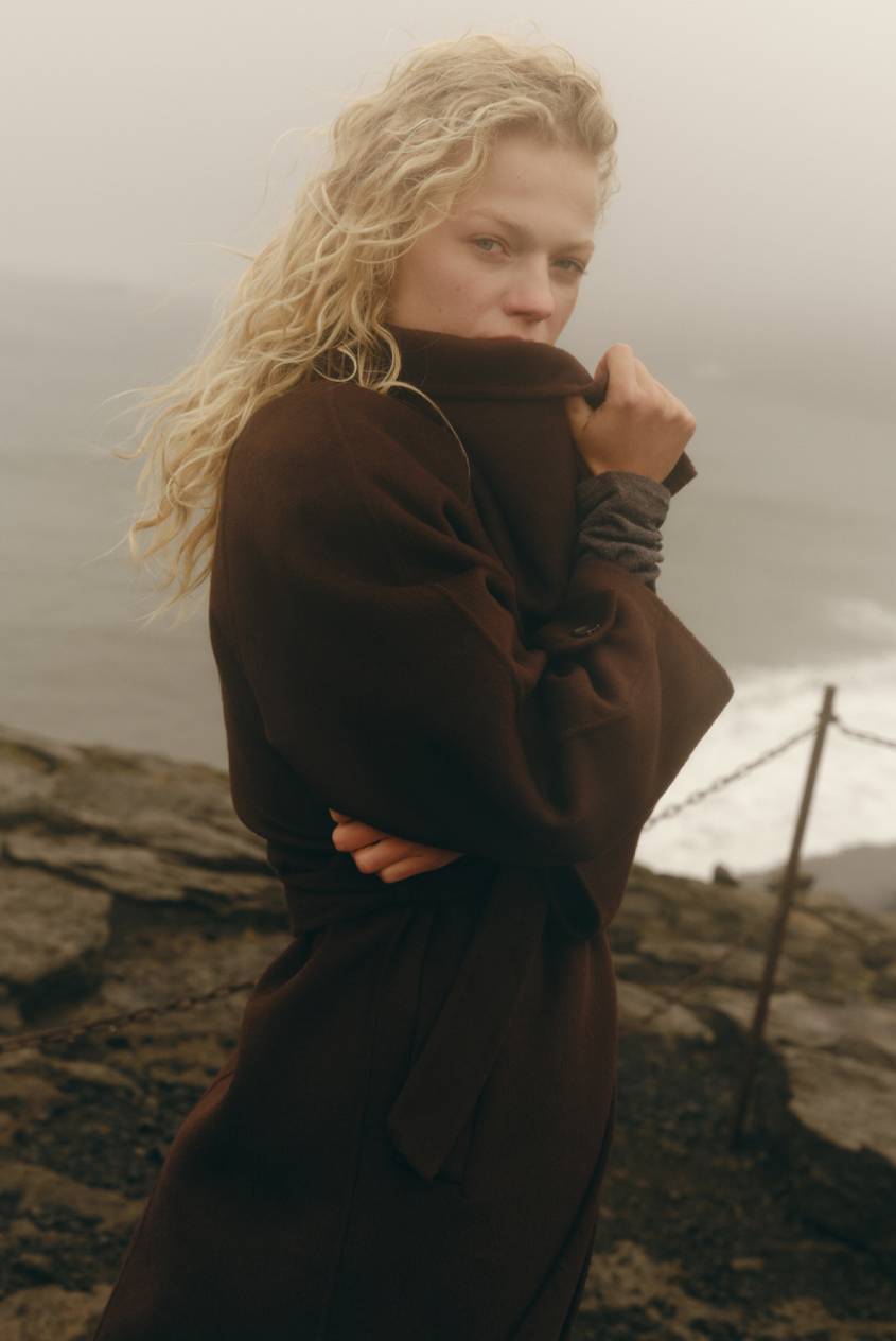 Blonde model wearing a dark brown coat standing on a rocky coastline with the ocean in the background.