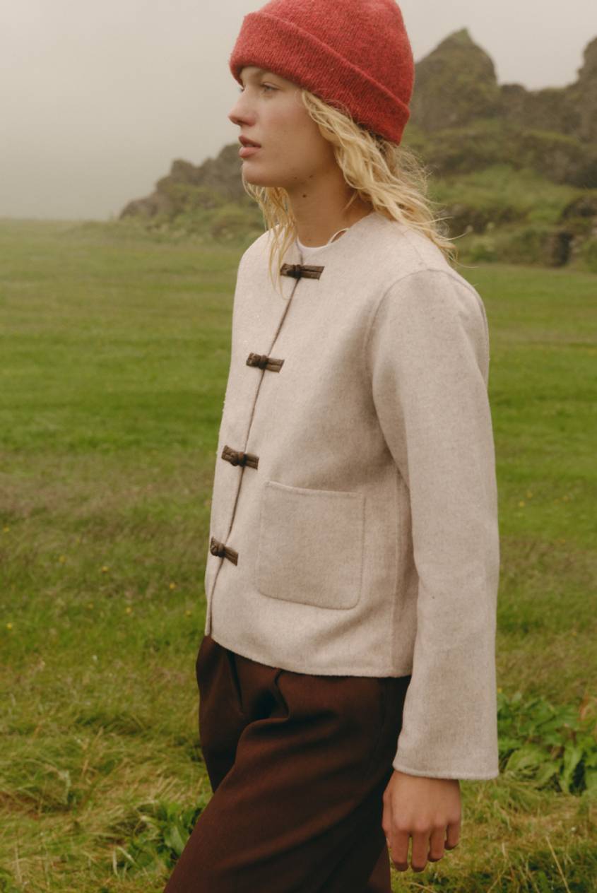 Blonde model wearing a red beanie, cream toggle-front jacket, and brown pants standing in a grassy landscape.