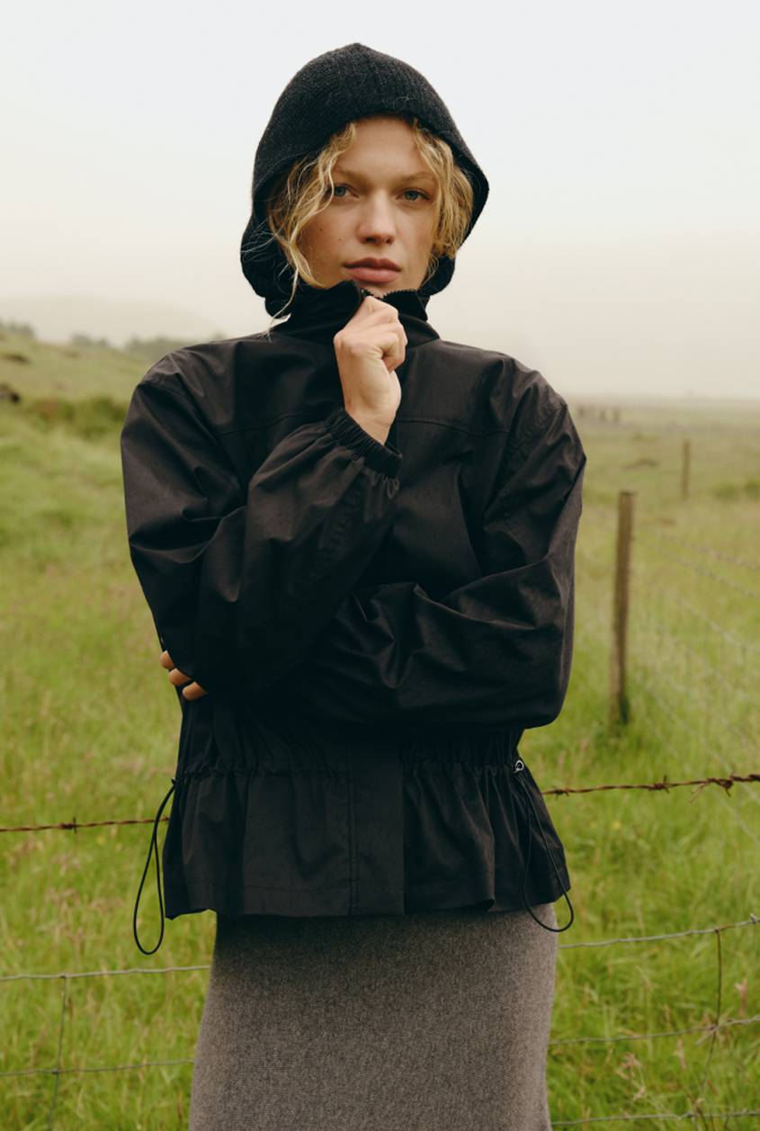 Model wearing a black hooded jacket and gray skirt standing in a grassy field with a fence in the background.