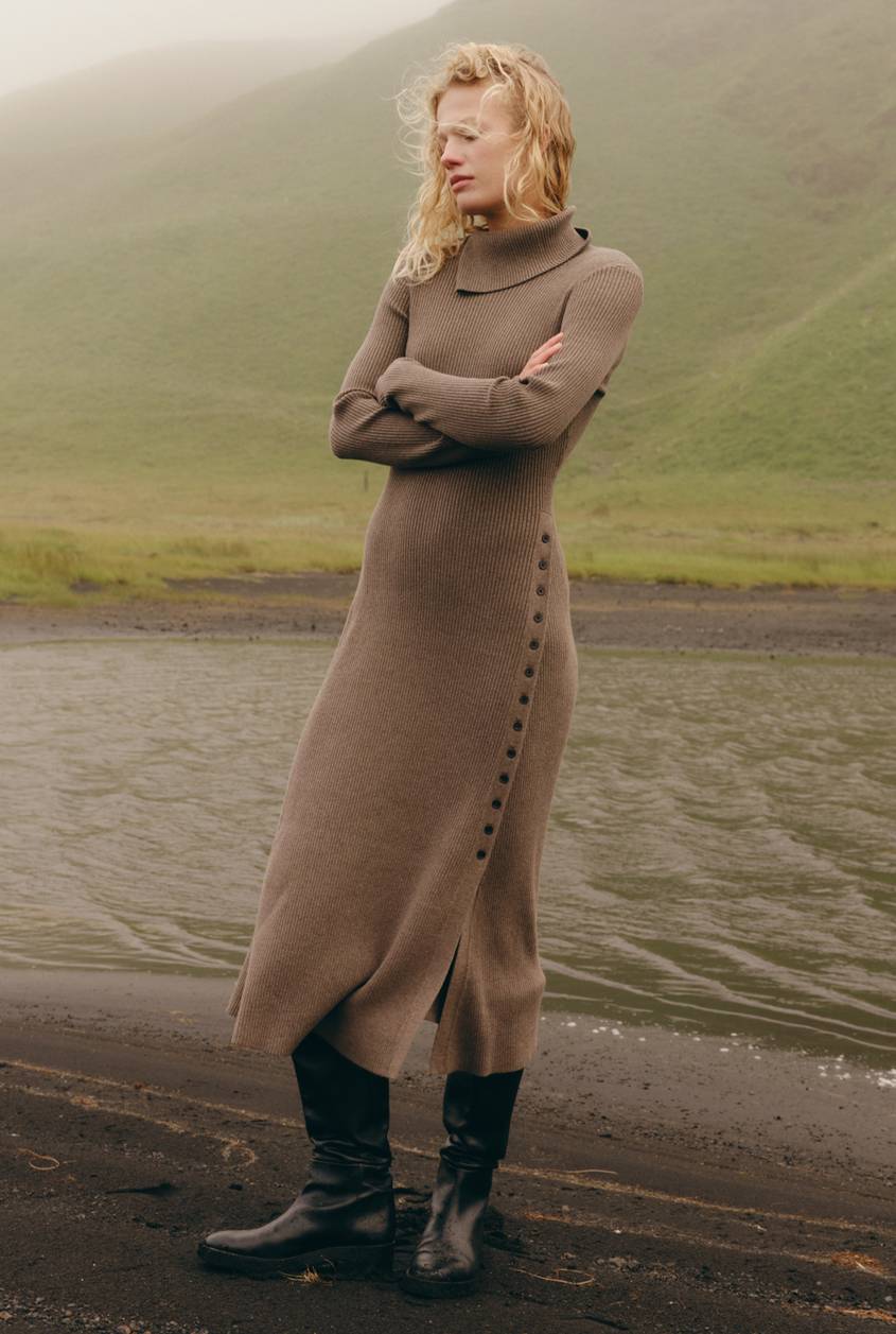 Model in a taupe ribbed turtleneck sweater dress with a side button placket and black knee-high boots, standing on a dark sand beach with arms crossed against a foggy green hillside backdrop.